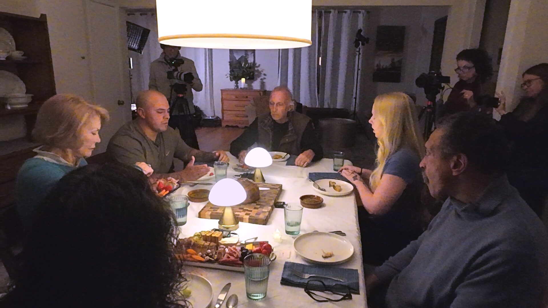 Six people sharing a meal at a lit table while the film crew works in the background