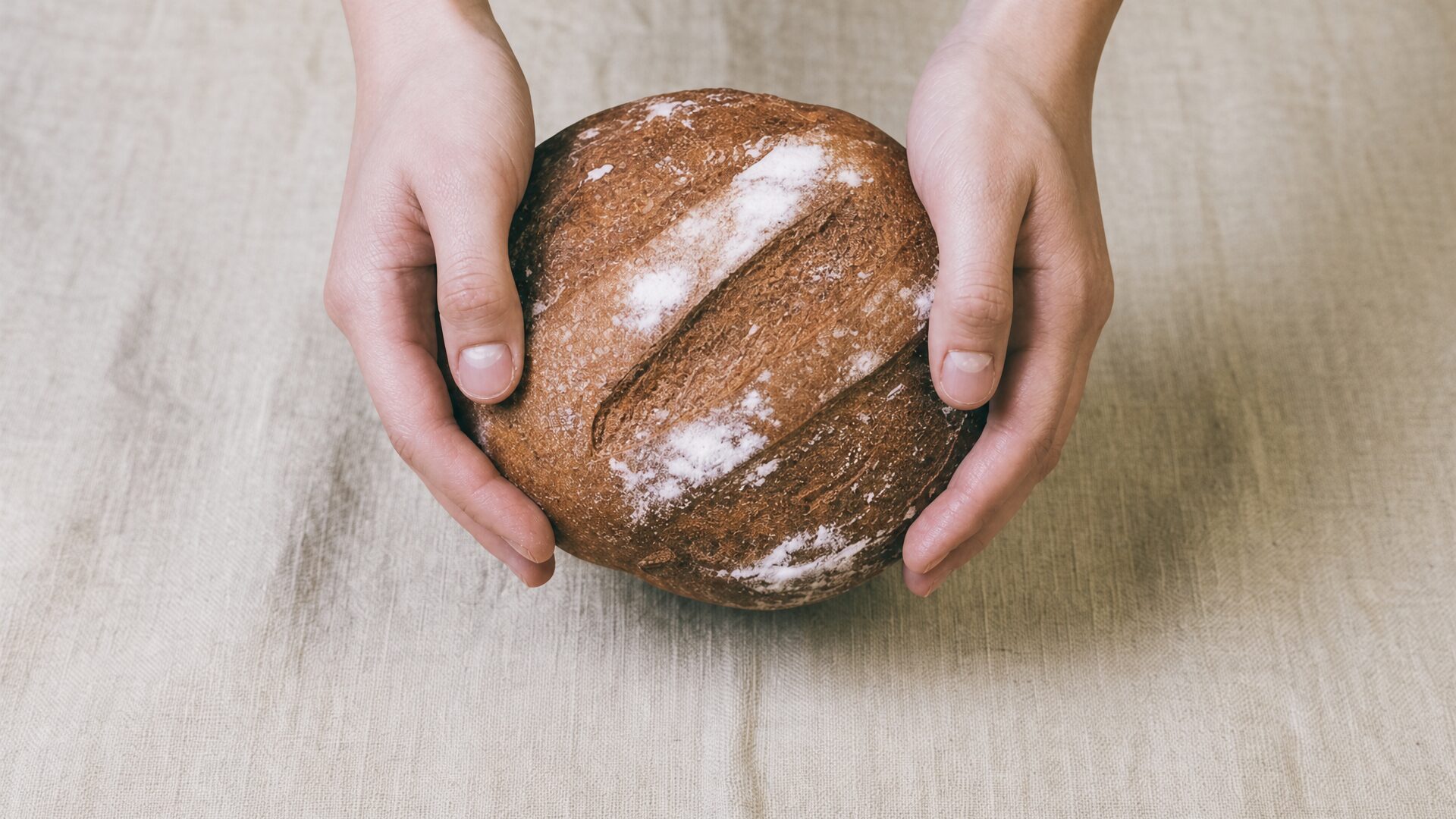Two hands holding a round flour-dusted loaf of bread over a linen tablecloth